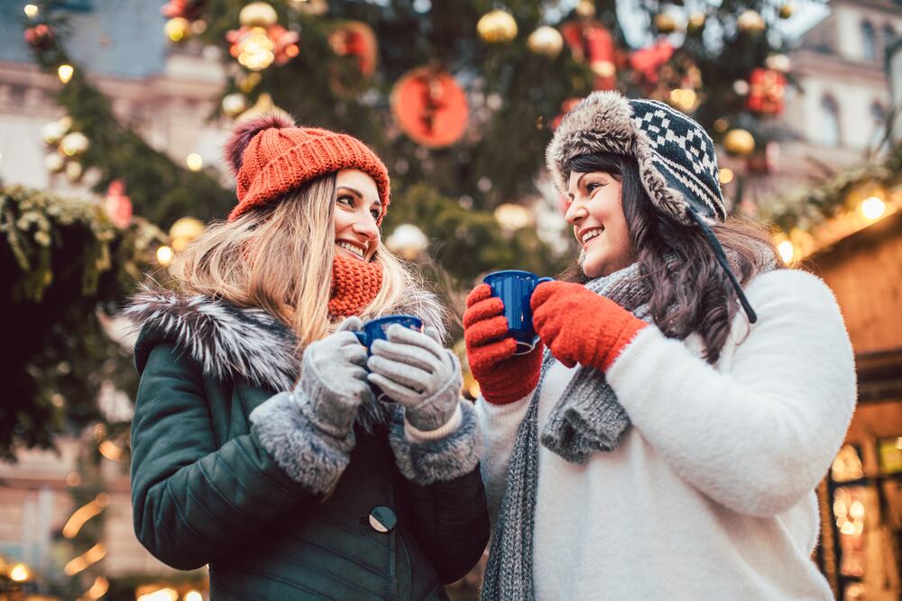 Zwei Freundinnen mit Wintermützen und Tassen lachen vor einem geschmückten Weihnachtsbaum