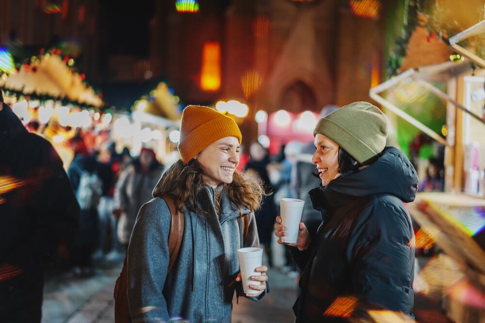 Zwei Frauen mit Wintermützen unterhalten sich lachend mit heißen Getränken zwischen Marktständen